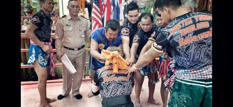 Wai Kru Mongkon ceremony in Thailand: Master Toddy and his instructors placing the Mongkon headband on our head instructor, kneeling, in front of Thai, Canadian and other national flags.