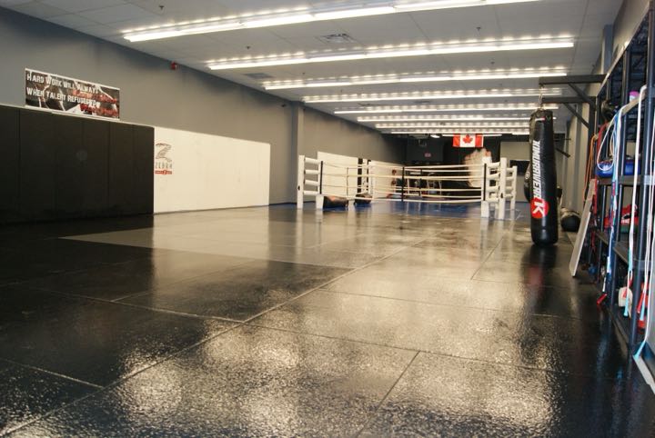 Inside Rumble Martial Arts: black mats, a white boxing ring, a heavy bag and a Canadian flag under a banner that reads Hard work will always beat talent when talent refuses to work hard.